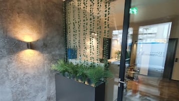 A modern lobby with a glass door, a planter with green plants, a textured wall, and a wall-mounted light fixture.