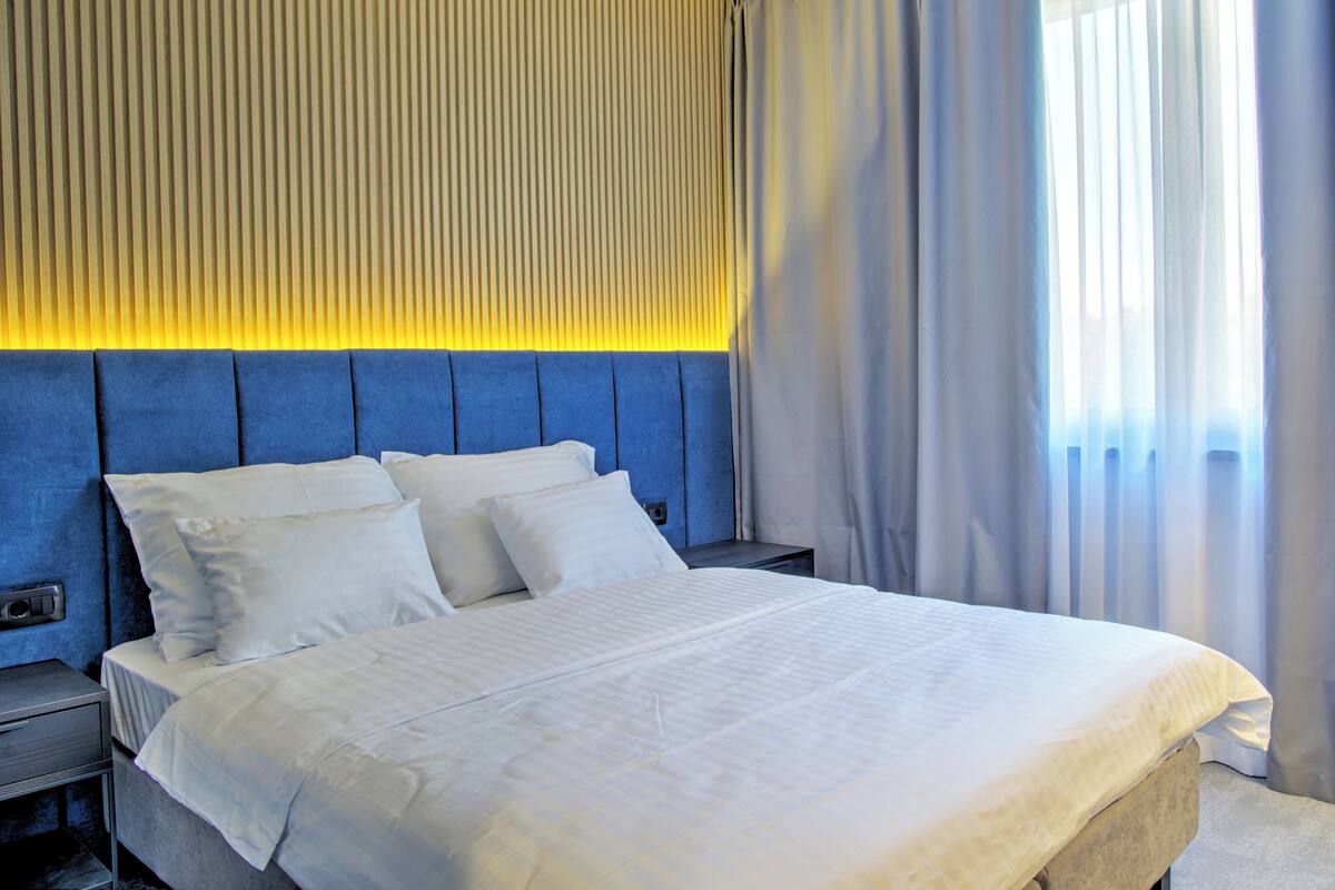 Una habitación de hotel con una cama grande, cabecera con líneas verticales, mesita de noche y ventana con cortinas.