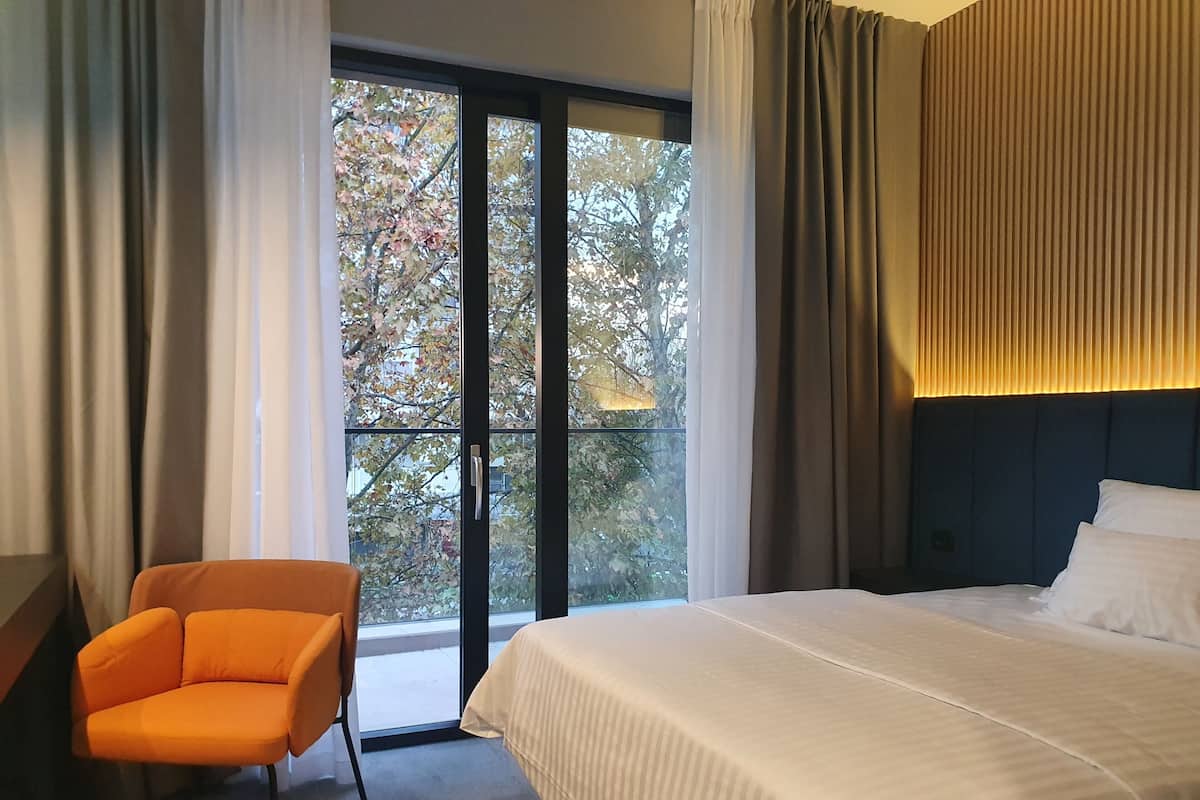 Una habitación de hotel con una cama, un sillón naranja y un ventanal con vistas a árboles.