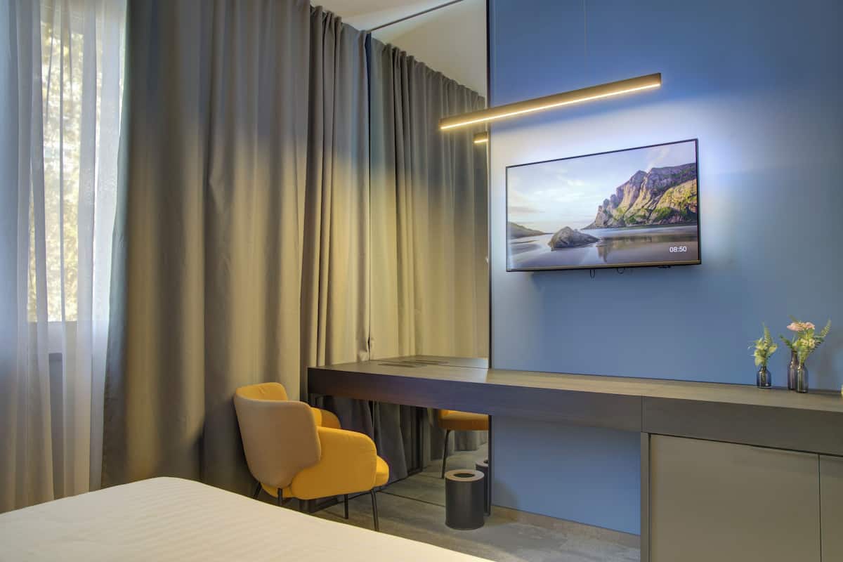 Una habitación de hotel moderna con una cama, un escritorio con una silla, un televisor montado en la pared y una ventana con cortinas.
