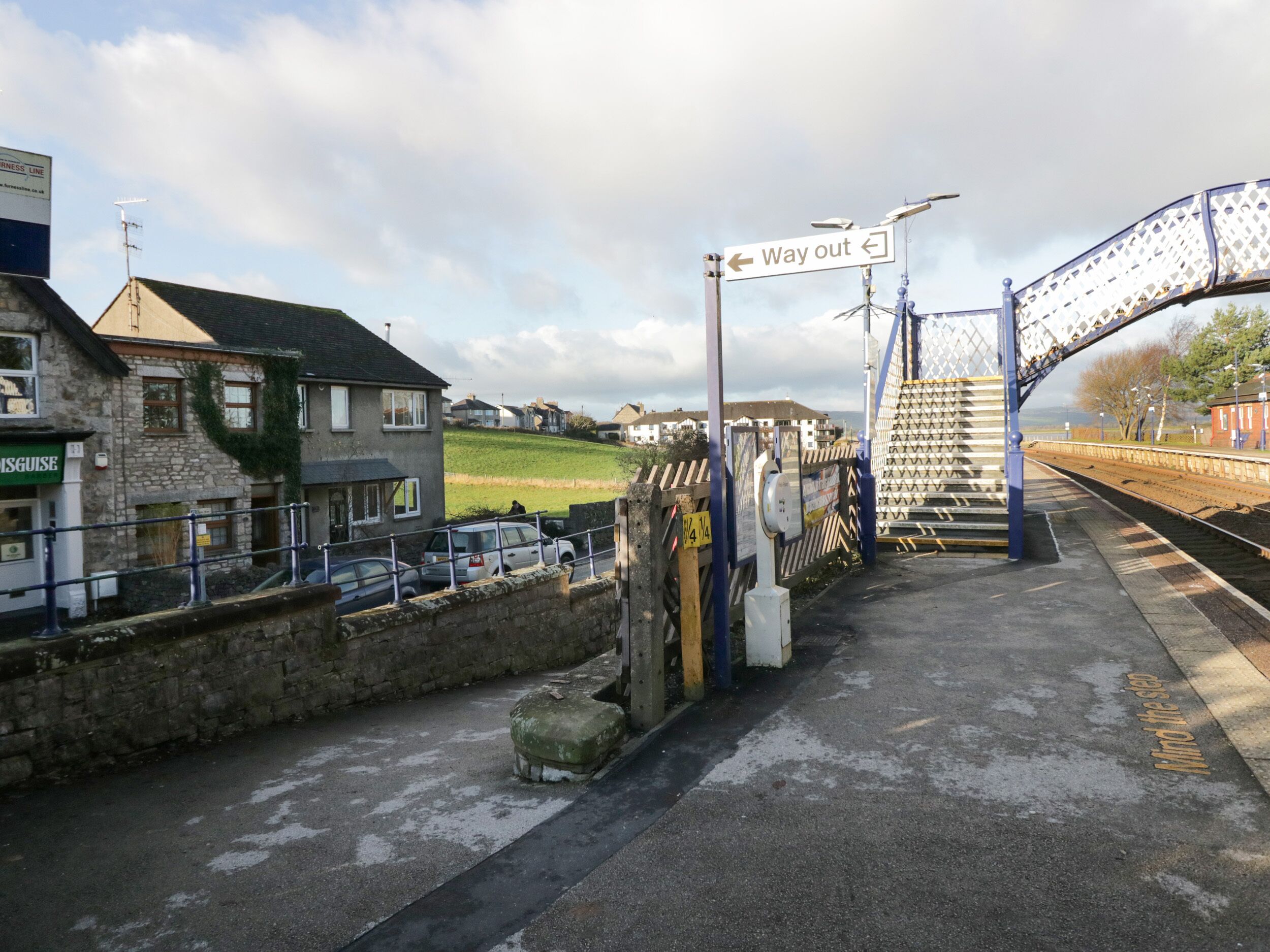 A railway station platform with a 'Way out' sign, a stone building with a green sign, and a bridge.