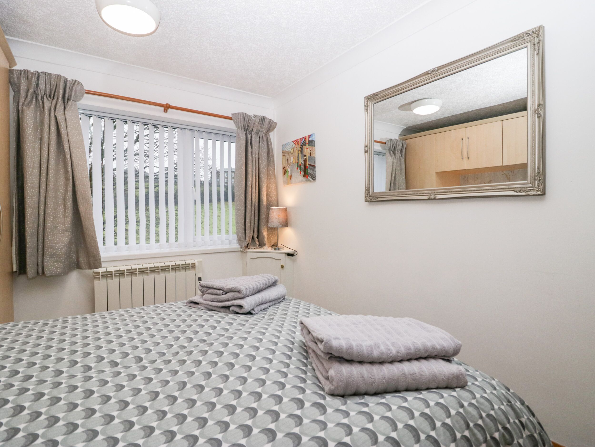 A bedroom with a bed, a window with curtains, a radiator, a mirror, and a wall-mounted lamp.