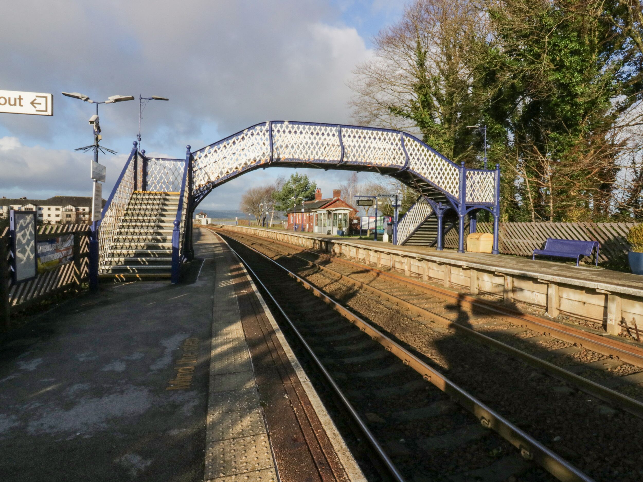 Un andén de estación de tren con un puente peatonal, vías y un edificio pequeño al fondo.