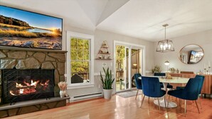 A living room with a fireplace, a dining area with blue chairs, and a large TV mounted on the wall. - The Outpost Camelback: Walk to Ski, Swim, Fun! (Tannersville)