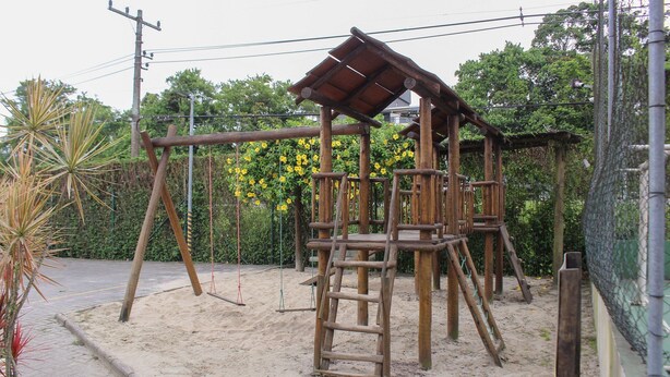 Une structure de jeu en bois, dotée d’une échelle et de balançoires, installée dans un espace sablonneux, entourée de verdure et d’une clôture.