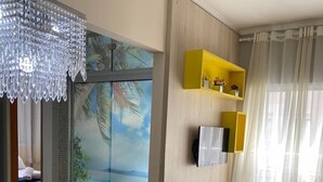 Un salon moderne avec un plancher en bois, un grand téléviseur fixé à un mur en bois, un lustre et un tableau représentant une scène tropicale.