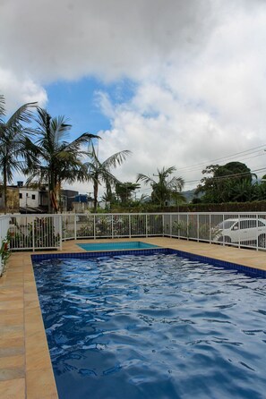 Une piscine dont le bord est recouvert de carrelage, entourée d’une clôture blanche, de palmiers et de bâtiments en arrière-plan.
