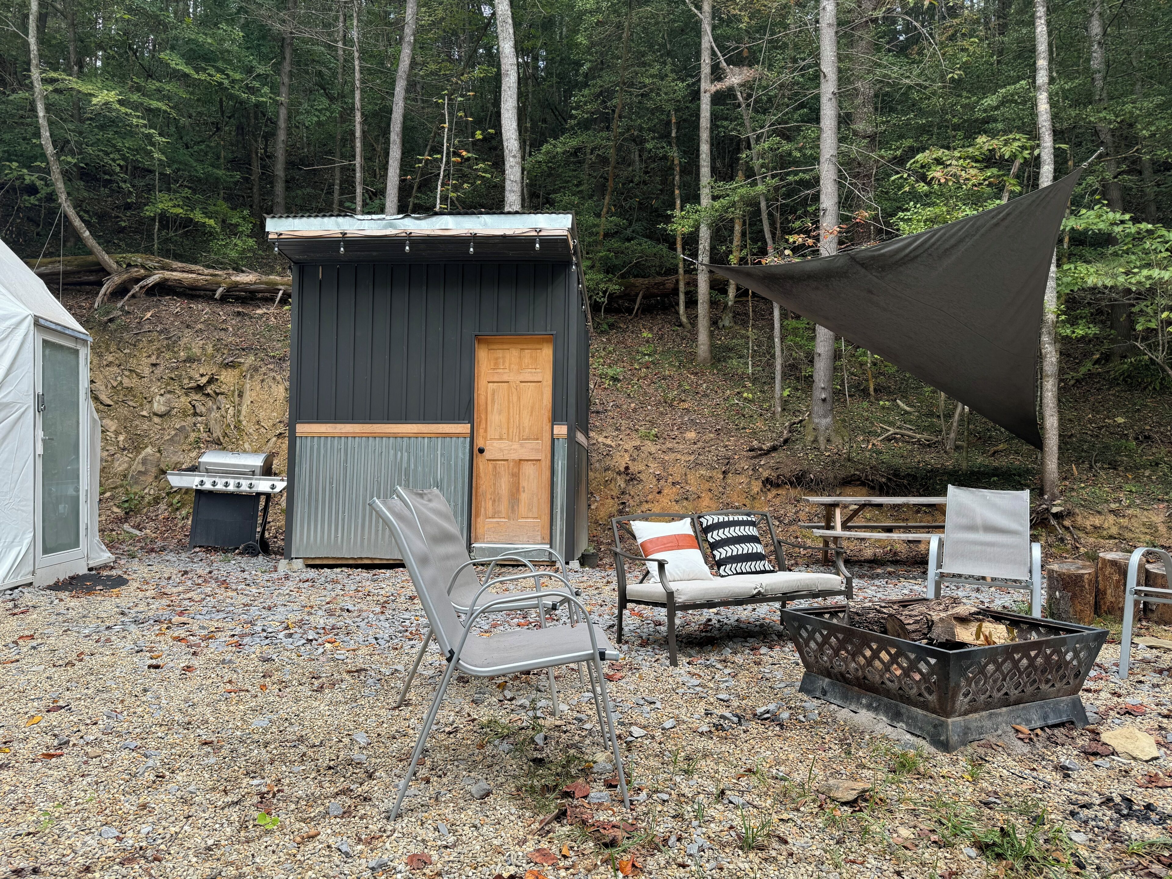 Eine rustikale Holzhütte mit Tür, Grill, Feuerstelle und Sitzgelegenheiten im Freien.
