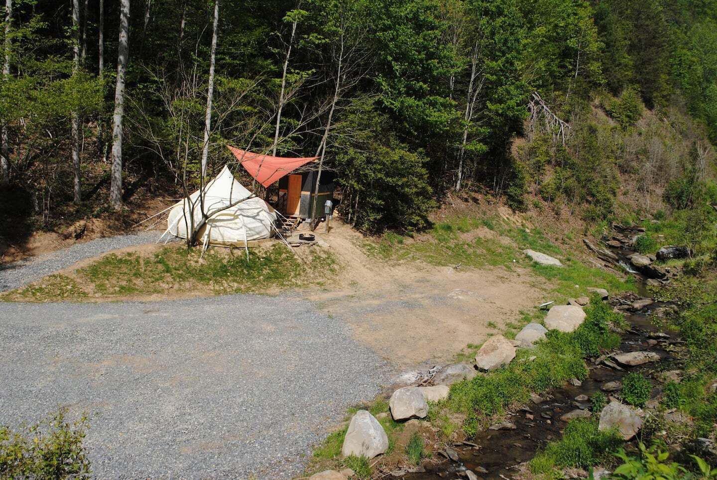 Ein Zeltplatz mit einem Kiesweg, der zu einem kleinen Gebäude führt, umgeben von dichtem Wald und einem Bach.