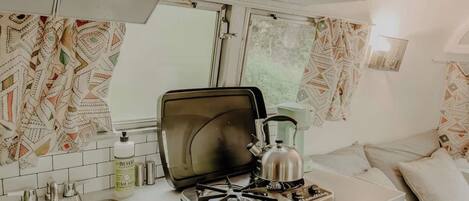 A compact kitchen with a gas stove, a kettle, and a coffee maker. A patterned curtain hangs over the window, and a shelf above the counter holds a few items.