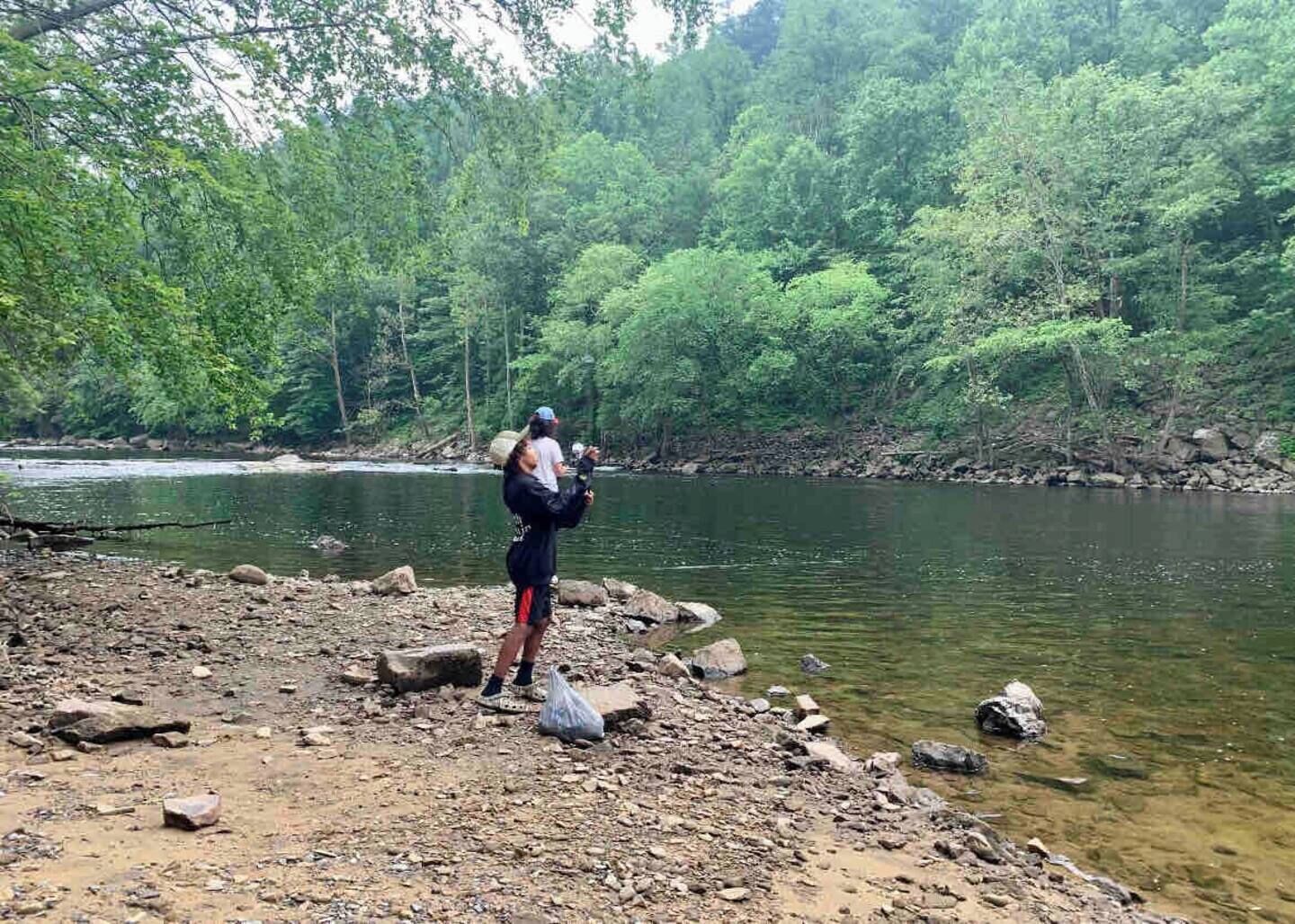 Une personne se tient sur la rive rocheuse d’une rivière, un sac à dos sur le dos, entourée d’une forêt dense et verdoyante.