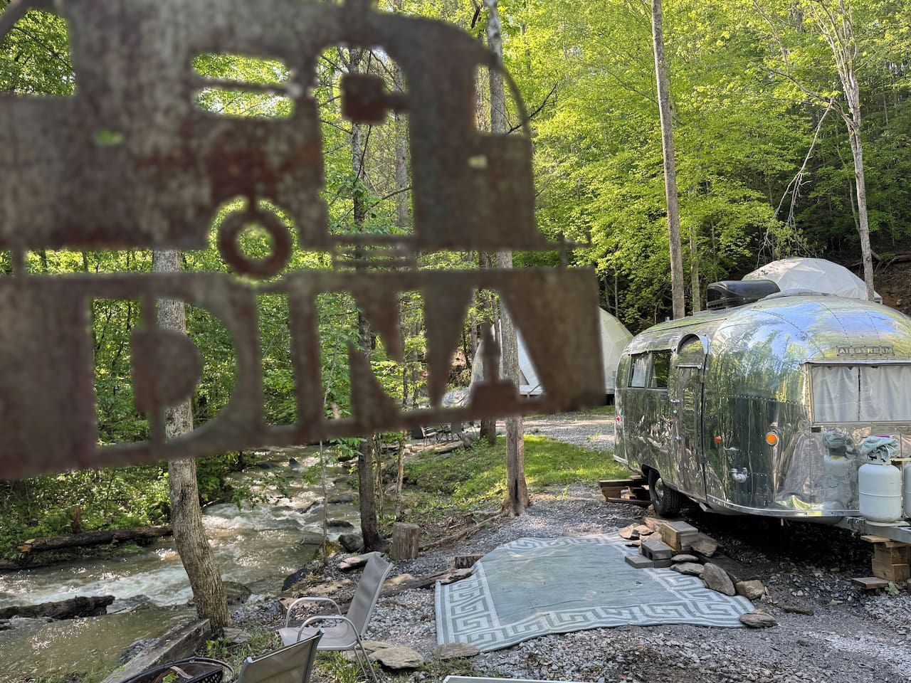 A vintage trailer parked in a forested area with a stream nearby.