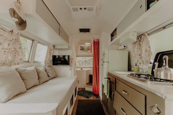 Interior compacto de una caravana con cocina americana, baño pequeño, zona de estar y televisor.