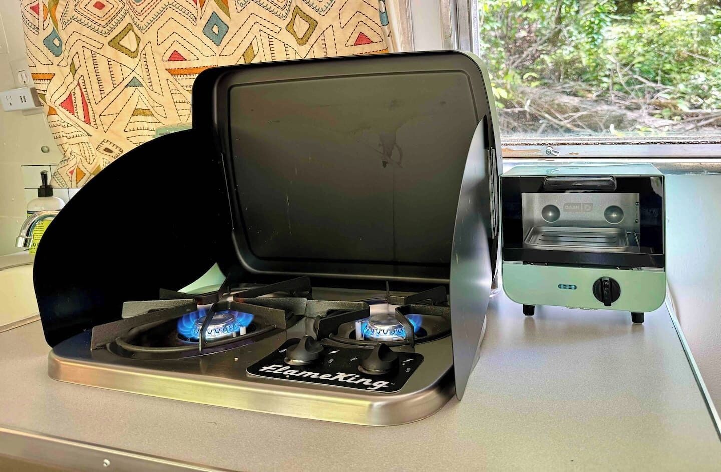 A portable gas stove with two burners and a small electric heater on a countertop.