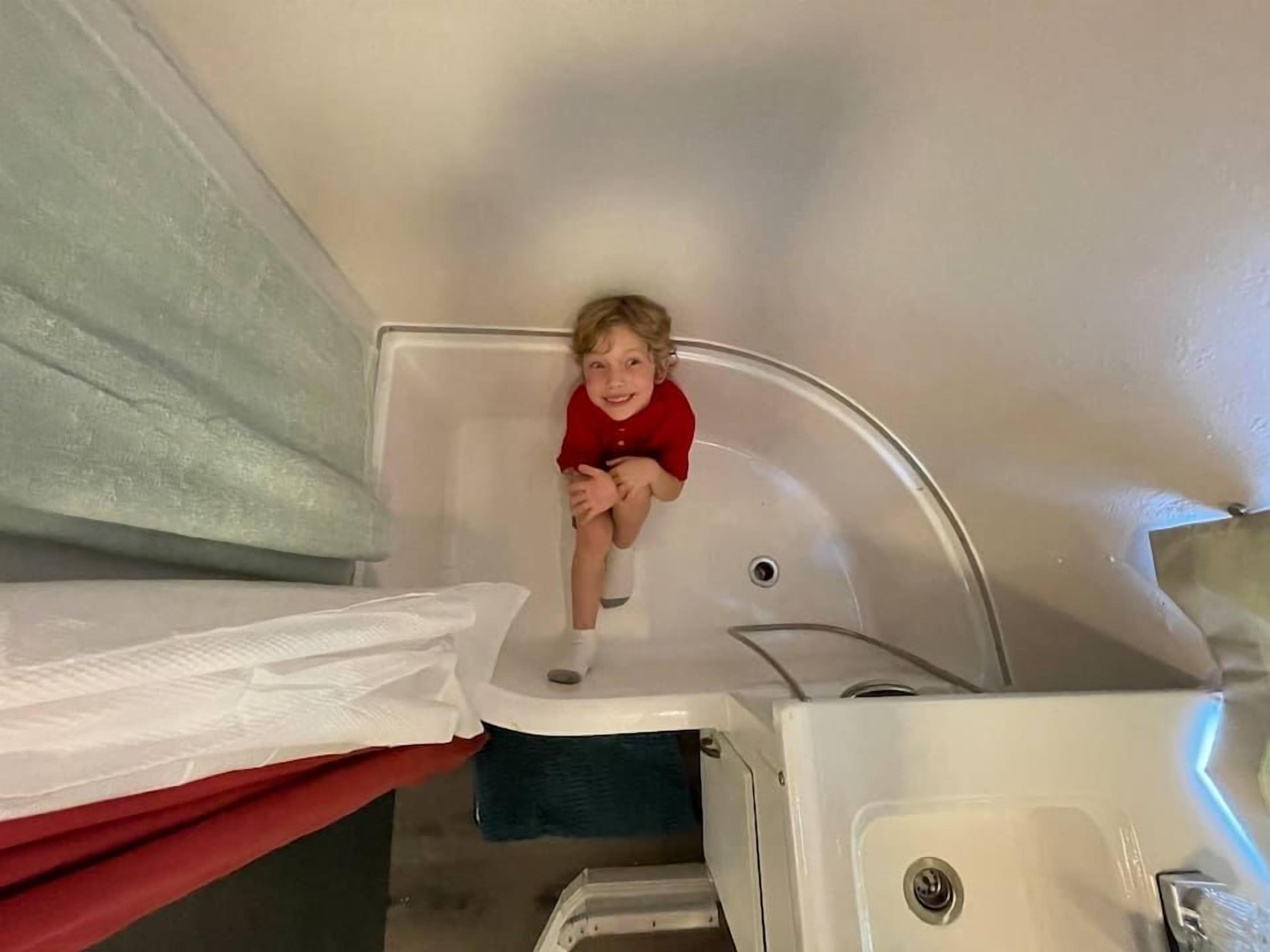 A child in a bathtub with a red shirt, a white sink, and a towel rack.