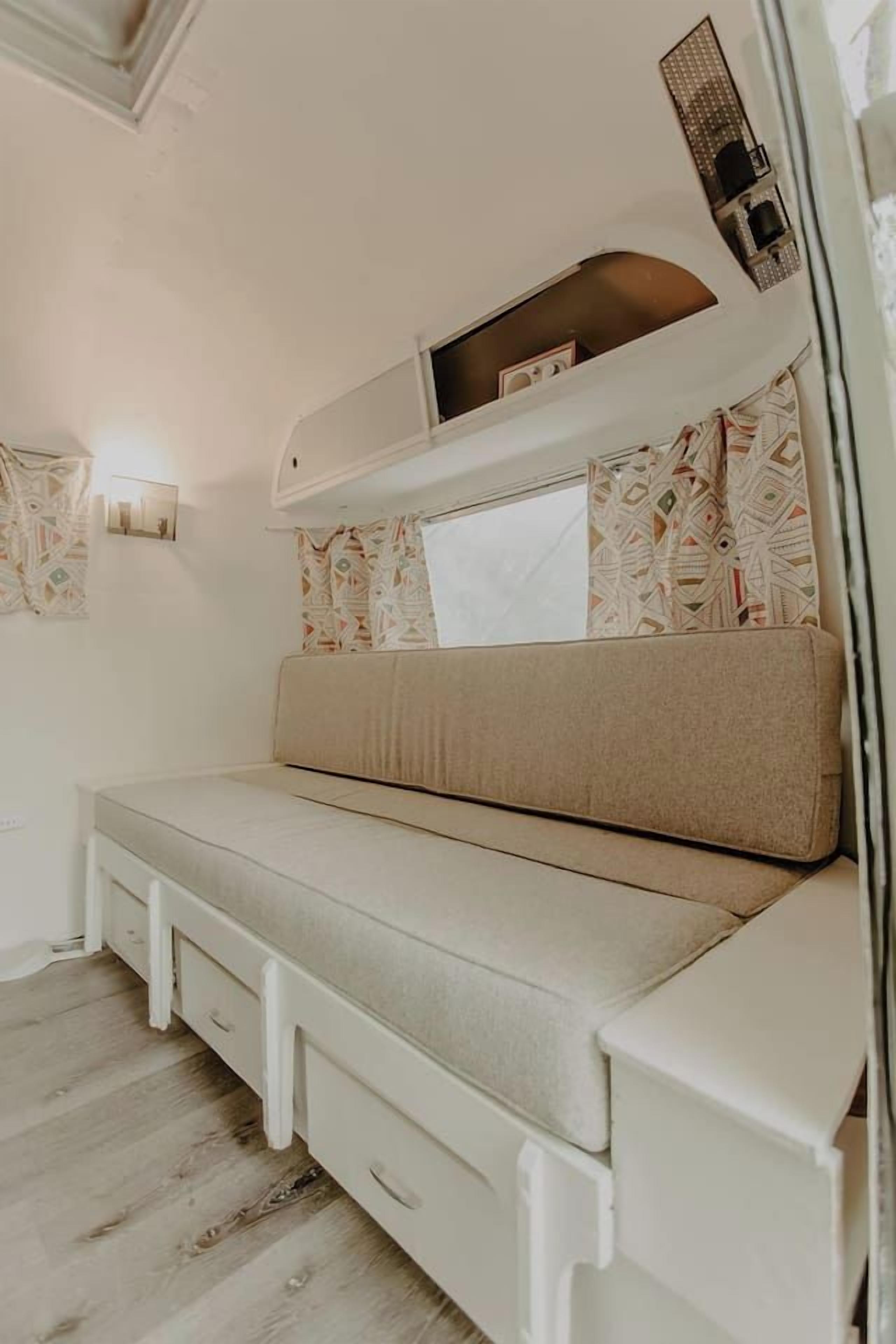 A compact, white interior with a sofa bed, a wall-mounted light, and patterned curtains.