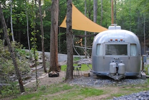 Un camping-car argenté est garé dans une zone boisée, sous un auvent jaune, entouré d’arbres et d’une petite table avec des chaises.
