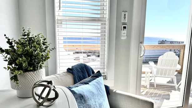 Un balcon donnant sur l’océan, une plante en pot blanche et un coussin bleu sur un canapé gris.