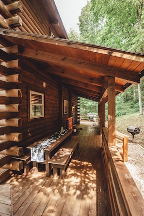 Relax on the covered porch with a wooden bench and table, surrounded by lush greenery.
