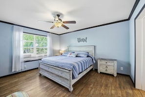 A bedroom with a bed, a nightstand, a ceiling fan, a window with curtains, and a wall sign.