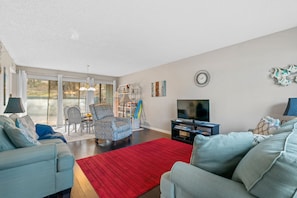 A living room with a red rug, two light blue sofas, a patterned armchair, a dining table with chairs, a bookshelf, a TV, and a large window with curtains.
