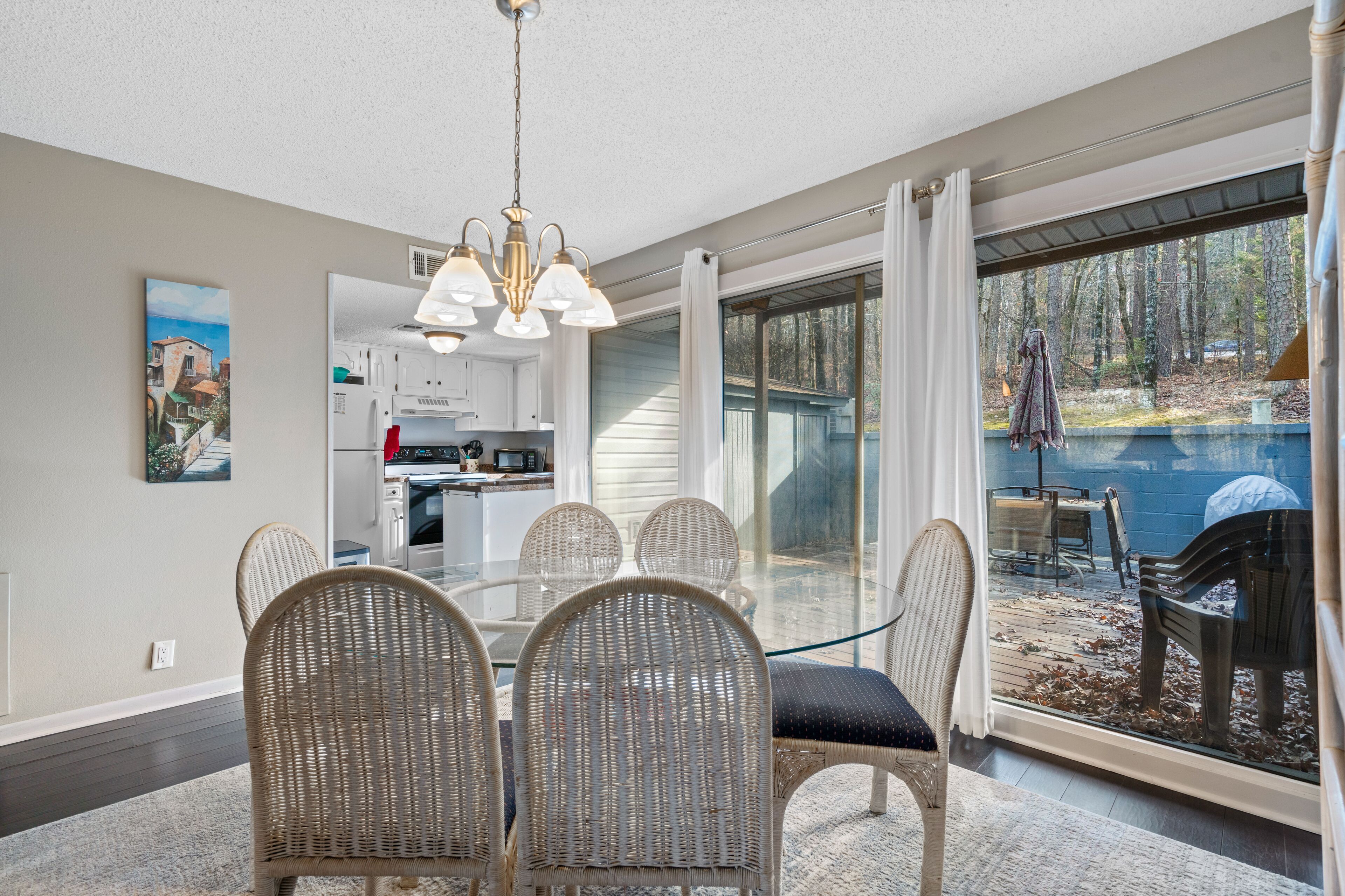 Un coin repas avec des chaises en osier et une table en verre, une cuisine à l’arrière-plan et une vue sur un jardin avec une piscine et des arbres.