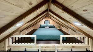 Un dormitorio tipo loft con techo inclinado, vigas de madera, una cama con colcha verde y un ventanal pequeño.