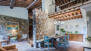 A rustic interior with a stone wall, wooden beams, a dining area with blue chairs, and a kitchen with wooden cabinets.