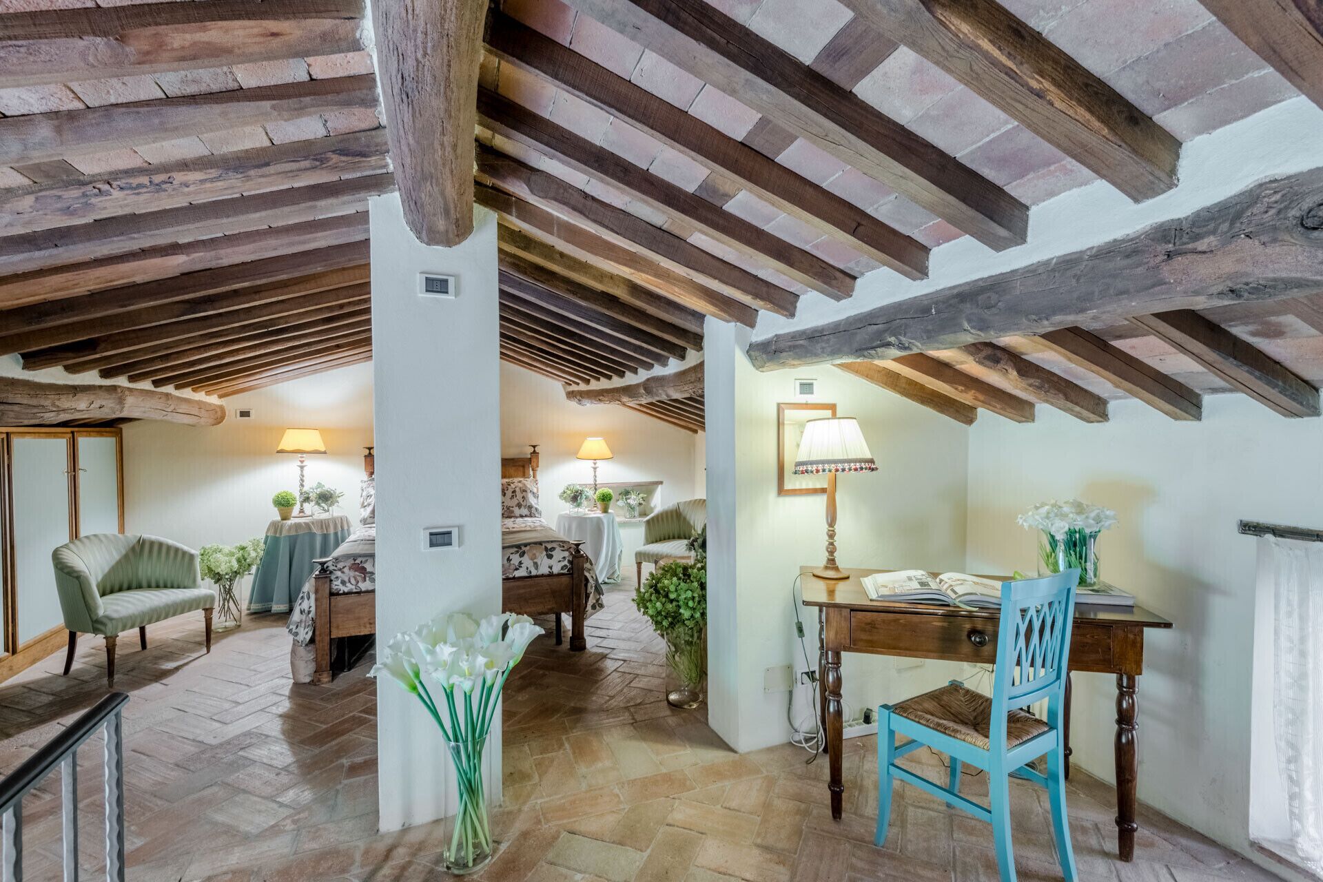 A rustic room with exposed wooden beams, a wooden desk, a chair, a bed, and a table with lamps.