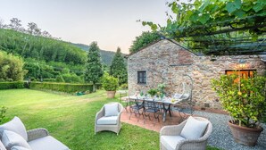 A stone building with a table and chairs set up outside, surrounded by a grassy yard and potted plants.