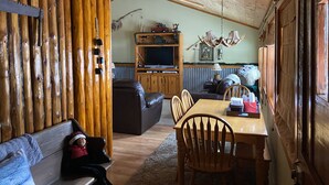Een gezellig chaletinterieur met een houten plafond, een televisie op een houten dressoir, een eettafel met stoelen en een bank met een kind erop.