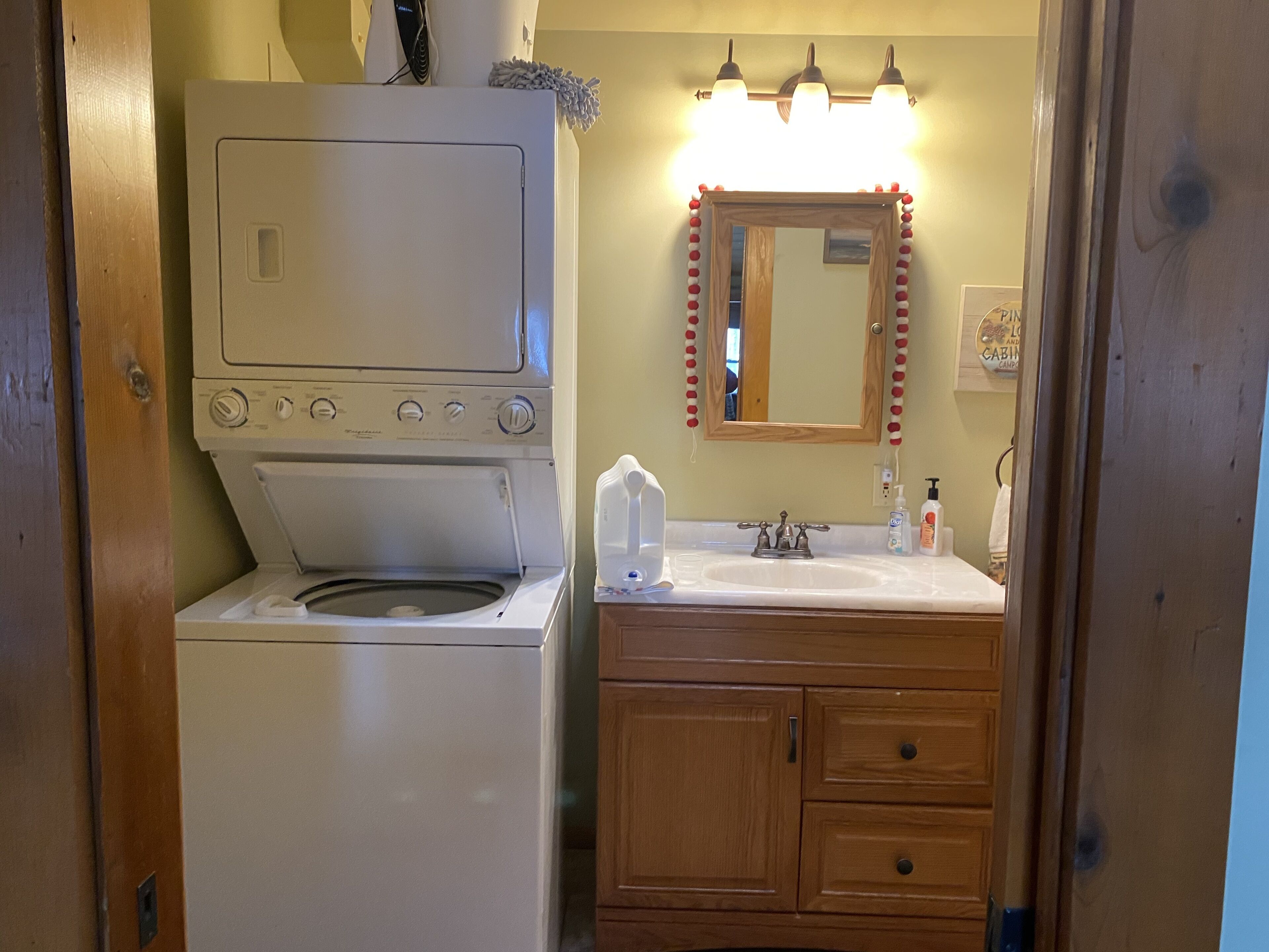A compact bathroom with a white washing machine, wooden cabinets, a sink with a mirror, and a light fixture above the mirror.