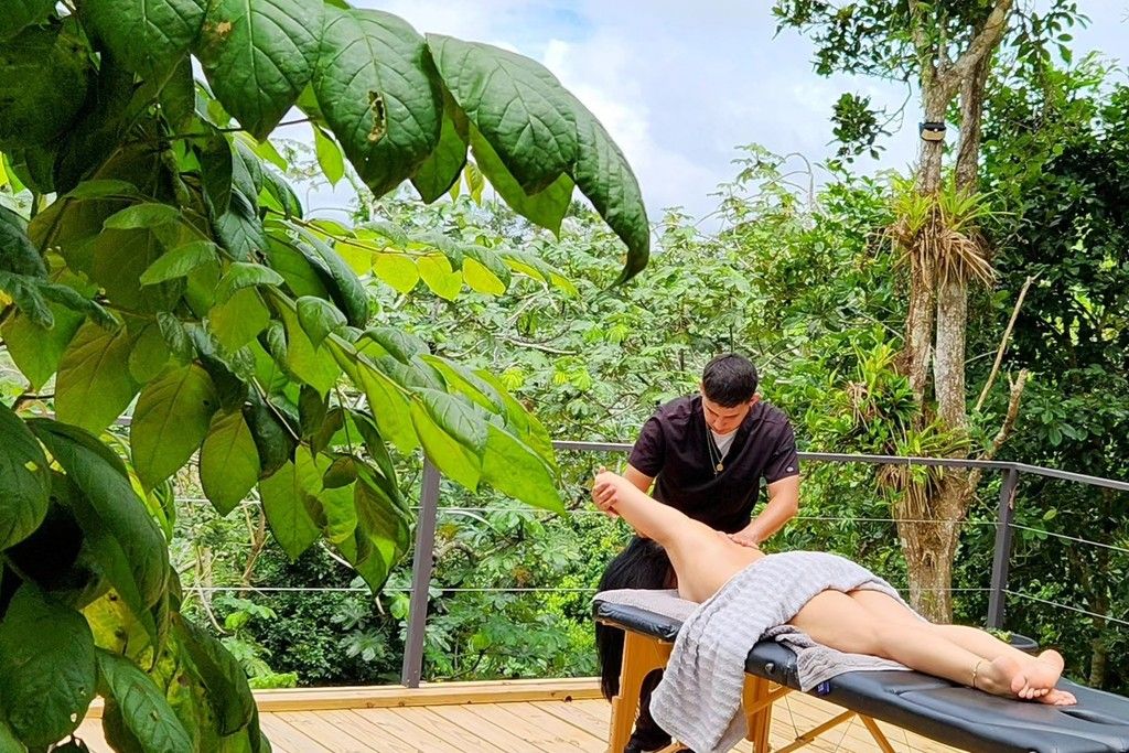 Uma pessoa recebendo massagem em um deck de madeira, com vegetação exuberante ao fundo.