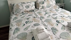 Een bed met een gewatteerde sprei met zee schelpen en koralen, een wit hoofdbord en een nachtkastje met een lamp en zakdoosjes.