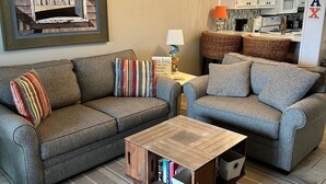 Een woonkamer met twee grijze banken, een houten salontafel, een lijst met een afbeelding en een keuken op de achtergrond.
