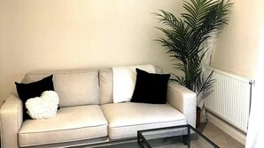 Een woonkamer met een beige bank, zwarte kussens, een glazen salontafel en een grote groene plant.