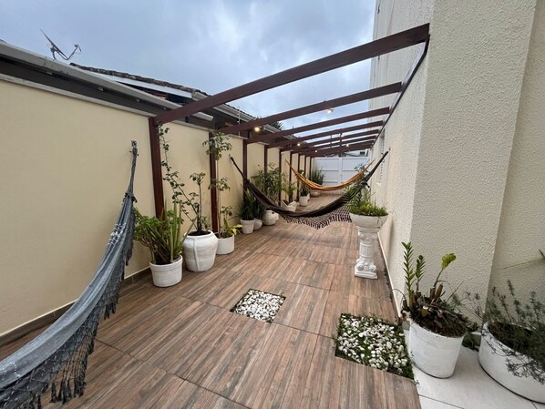 Un patio couvert avec un plancher en bois, des plantes en pot, un hamac et une vue sur un bâtiment en arrière-plan.