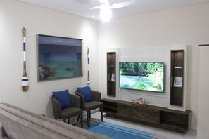Un salon avec un téléviseur à écran plat fixé au mur, un meuble de rangement en bois et deux fauteuils bleus rembourrés.