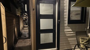 Un étroit sentier mène à une porte de couleur sombre, dotée de panneaux de verre dépoli, ainsi qu’à un petit espace extérieur aménagé avec une table et des chaises, et une fenêtre avec des stores se trouve sur la droite.