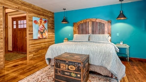 A bedroom with a wooden headboard, a bed with a patterned bedspread, a nightstand with a lamp, and a vintage suitcase. - The Red Barn Retreat, a reclaimed  grain barn centrally located in Kalispell MT (Kalispell)