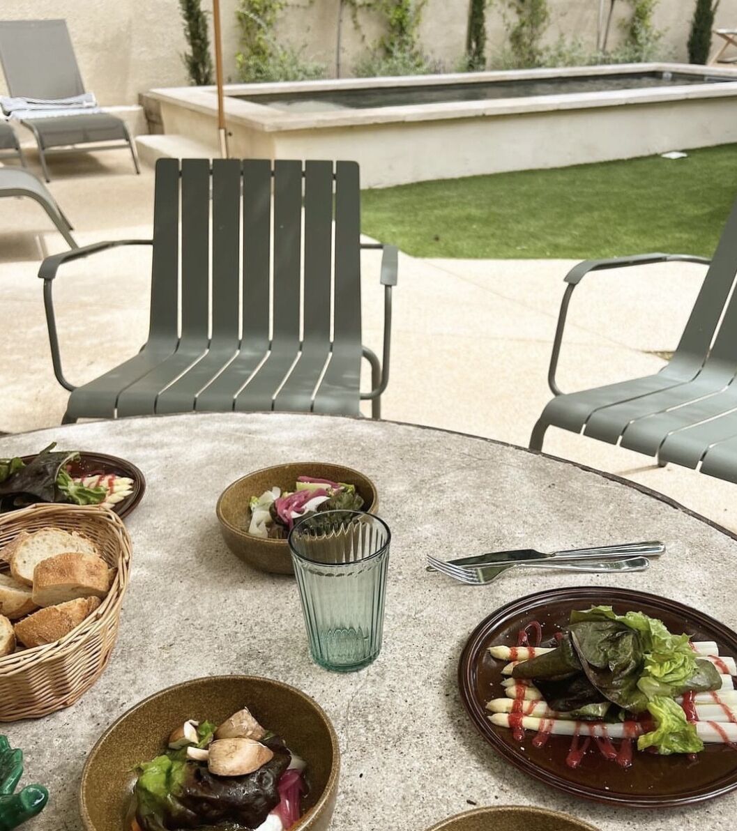 Ein runder Tisch mit verschiedenen Speisen, darunter Salate und Brot, auf einer Terrasse mit Stühlen und einem Rasen im Hintergrund.