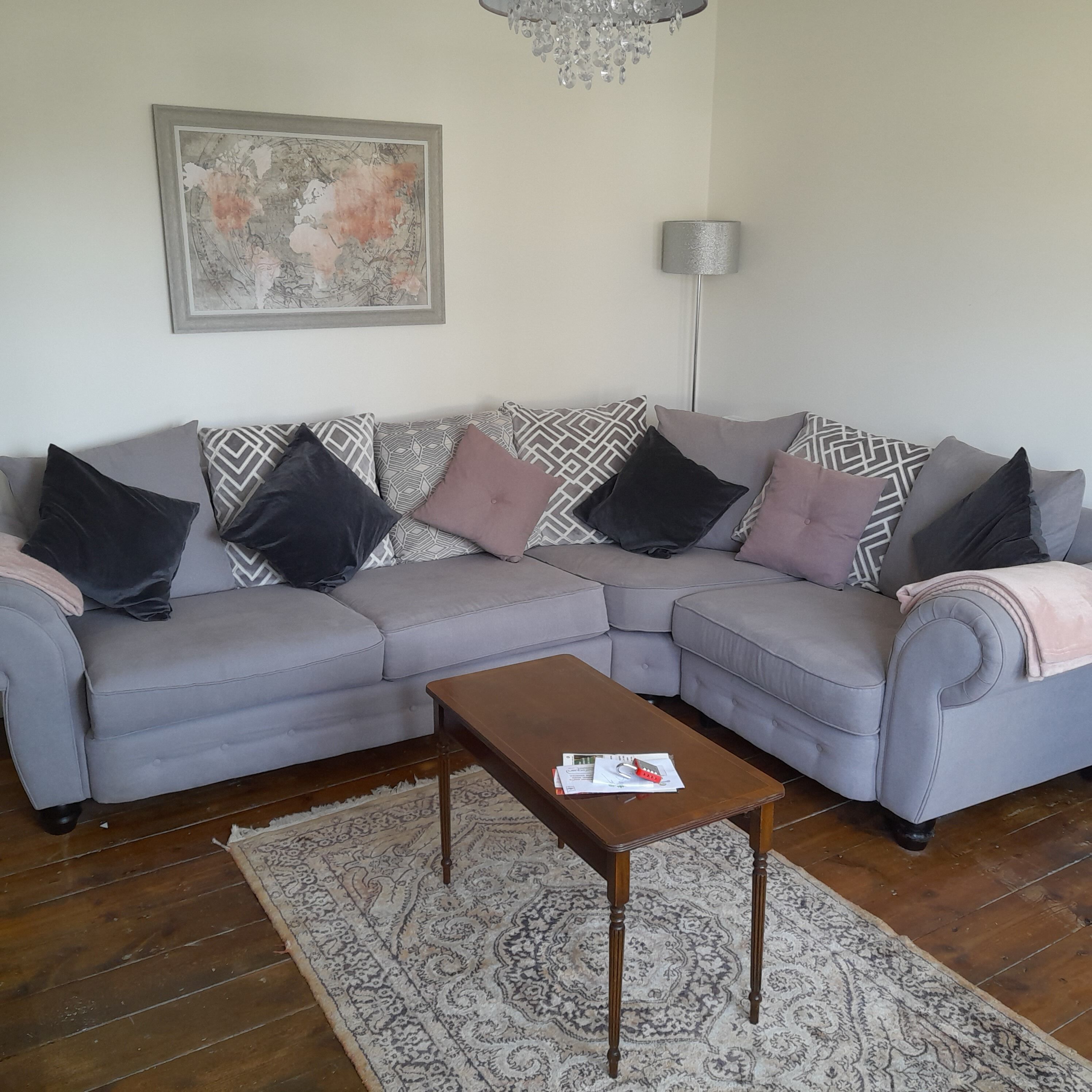 A living room with a grey sectional sofa, patterned cushions, a wooden coffee table, a patterned rug, a floor lamp, and a framed artwork on the wall.