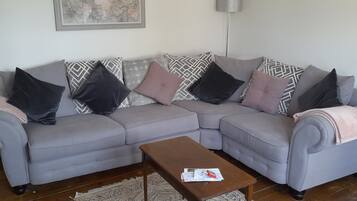 A living room with a grey sectional sofa, patterned cushions, a wooden coffee table, a patterned rug, a floor lamp, and a framed artwork on the wall.
