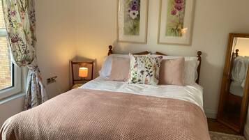 A neatly made bed with a pink blanket, a wooden headboard, and two floral pillows. There is a bedside lamp on a wooden nightstand, and two framed floral paintings on the wall.