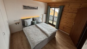 Een slaapkamer met een houten plafond, een bed met beddengoed met een patroon, een nachtkastje, een wandlamp en een raam met gordijnen.