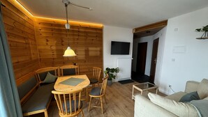 Une pièce chaleureuse avec un décor intérieur en bois, une table ronde en bois entourée de chaises, un canapé, un téléviseur fixé au mur et une plante en pot.