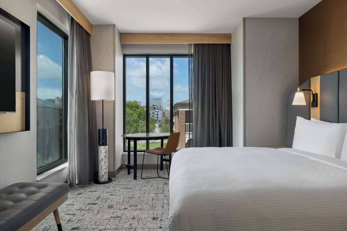 A hotel room with a large bed, a desk, and a chair. There is a window with a view of buildings and trees.