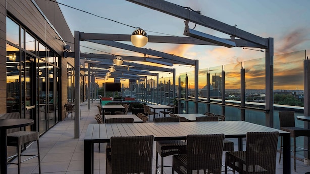 An outdoor terrace with tables and chairs, large windows, and a view of the city skyline at sunset. - Tempo By Hilton Louisville Downtown Nulu (Louisville)