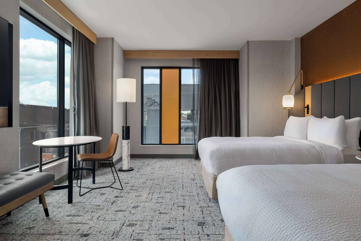 A hotel room with two beds, a small table with a chair, a sofa, a large window with curtains, and a view of the city.