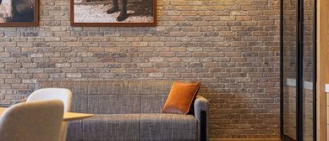 A cozy room with a brick wall, a grey sofa with a brown cushion, and a glass partition.